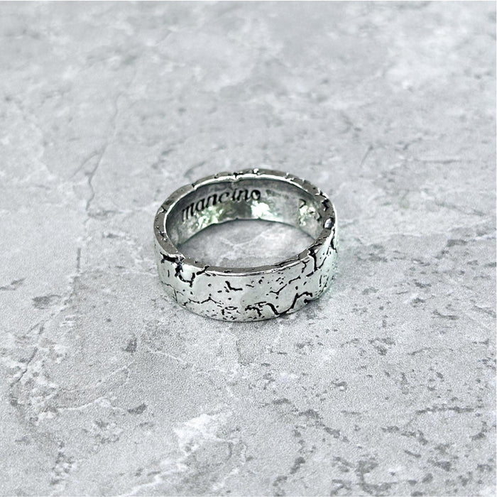 Nahaufnahme des Mancino "Broken" Rings aus 925 Sterling Silber mit markanter, rissiger Oberflächenstruktur und graviertem Logo auf der glatten Innenseite, liegend auf einem hellgrauen Steinuntergrund.