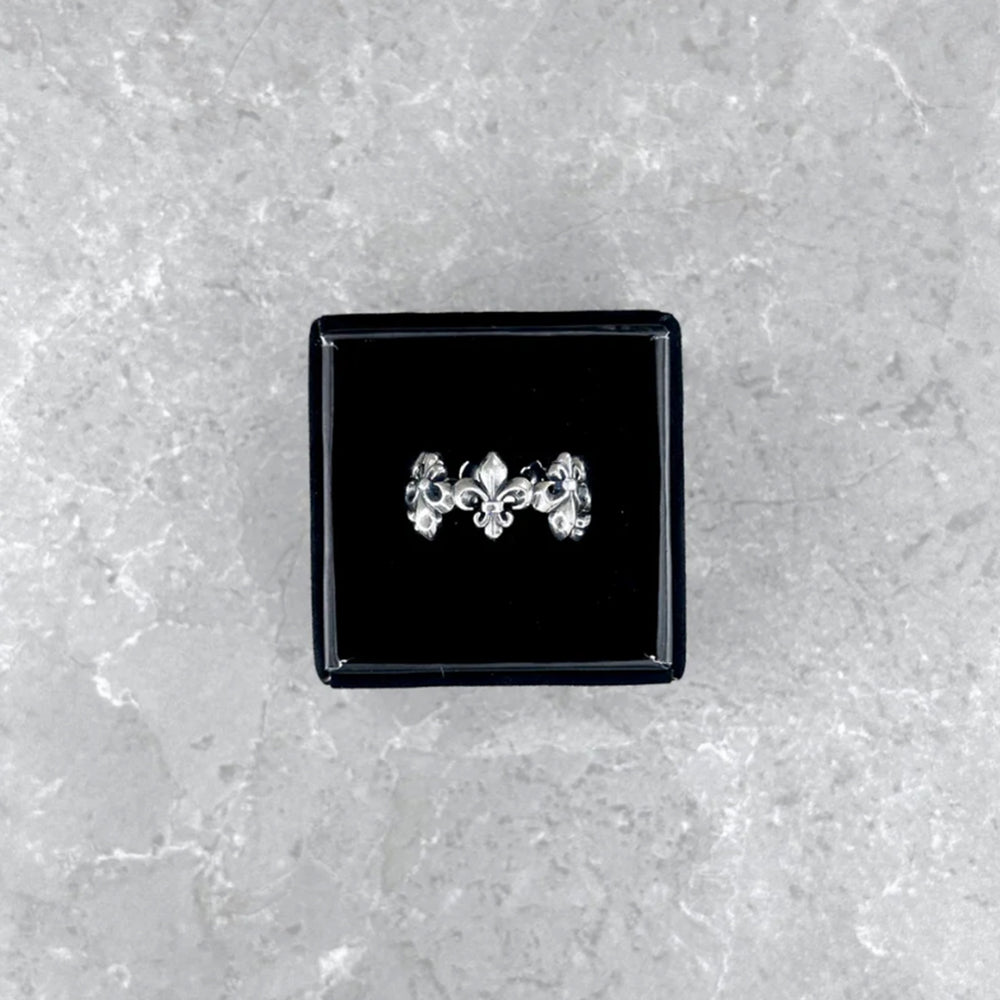 Der silberne Mancino "Giglio" Ring mit markantem Fleur-de-Lis-Muster, stilvoll präsentiert in einer geöffneten schwarzen Schmuckbox auf einem hellgrauen Steinuntergrund.