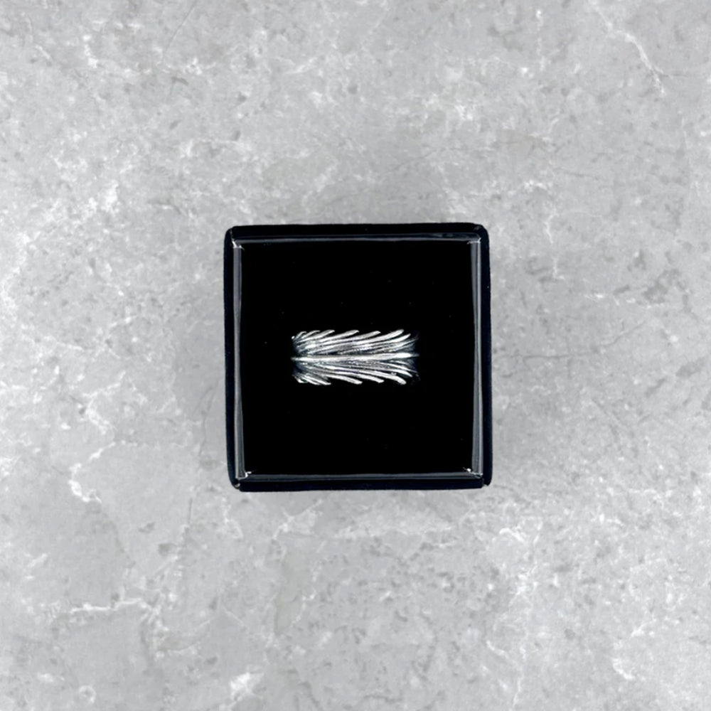 Der silberne Mancino "Piuma" Ring mit markantem Feder-Design, stilvoll präsentiert in einer geöffneten schwarzen Schmuckbox auf einem hellgrauen Steinuntergrund.