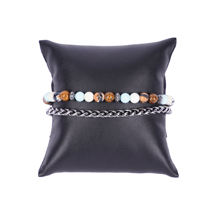 Zweireihiges Mancino Armband "Due Giri", elegant präsentiert auf einem schwarzen Kissen. Eine Reihe besteht aus einer massiven Edelstahlkette, die zweite aus 6mm Natursteinen in Braun, Weiß und Hellblau mit antiksibernen Metalldetails.