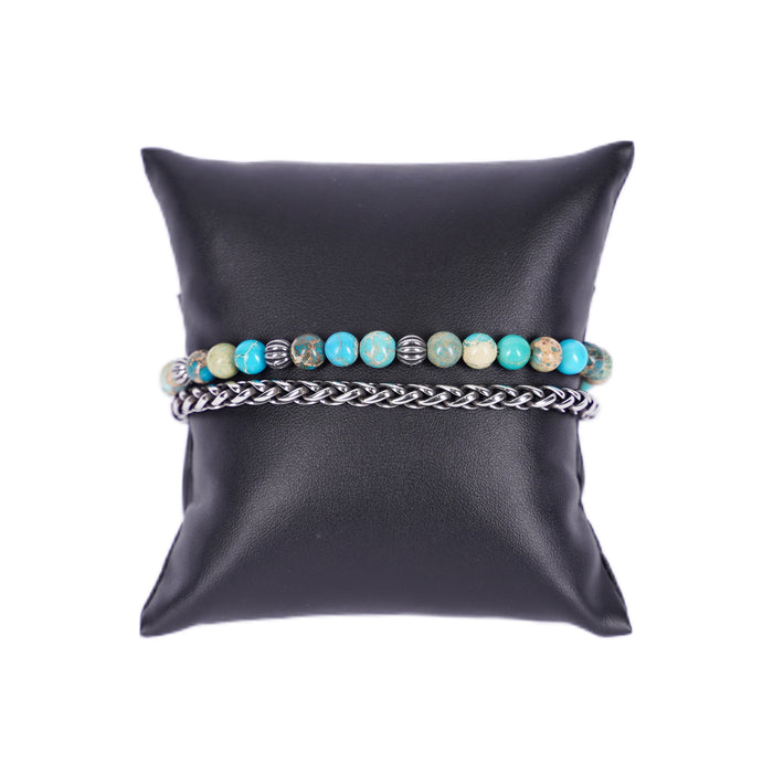 Zweireihiges Mancino Armband "Due Giri", elegant präsentiert auf einem schwarzen Kissen. Eine Reihe besteht aus einer geflochtenen Edelstahlkette, die zweite aus 6mm Natursteinen in leuchtendem Türkis mit erdigen Einschlüssen und antiksibernen Metalldetails.