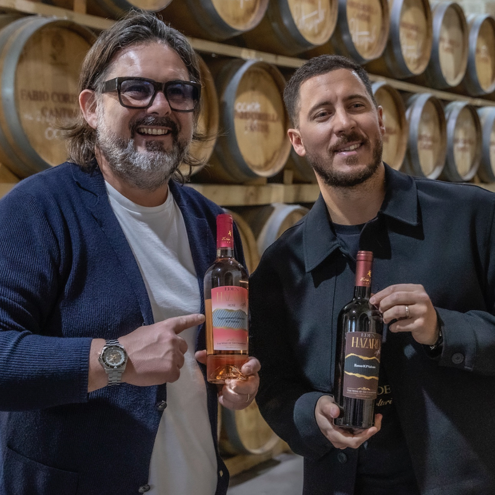 Fabio Cordella und die belgische Fußballlegende Eden Hazard stehen lächelnd in einem Weinkeller vor rustikalen Holzfässern. Beide präsentieren stolz die Weinflaschen aus der „Wine of the Champions“ Kollektion – Hazard hält den Rotwein, Cordella zeigt auf den Roséwein.