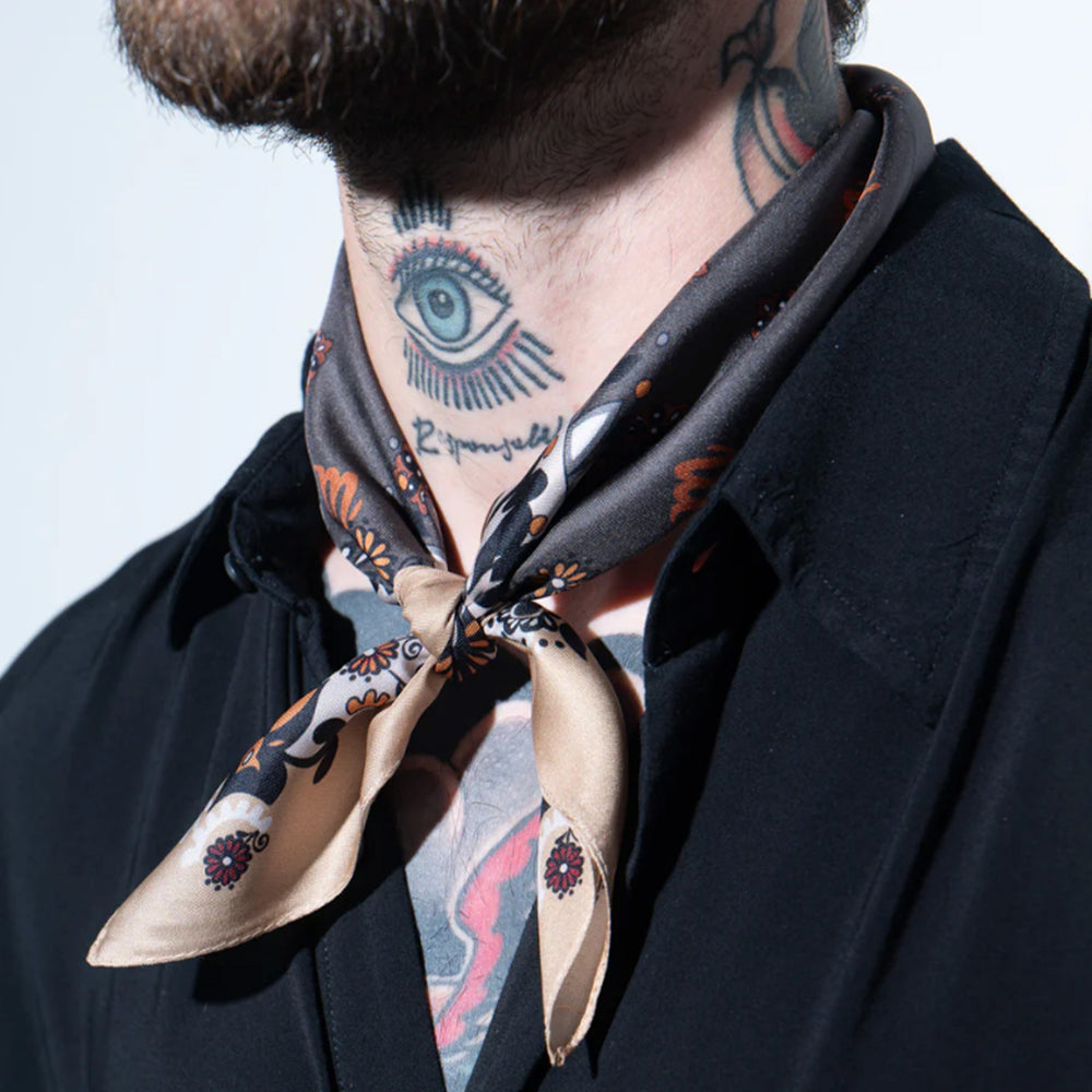 Nahaufnahme eines Mannes mit Tattoos am Hals, der das Mancino Seiden-Foulard locker über einem schwarzen Hemd geknotet trägt. Das Tuch zeigt edle Details in Sand, Orange und Dunkelgrau.