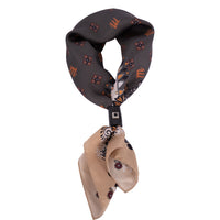 Mancino Seiden-Foulard (51x51 cm) in Dunkelgrau und Sand mit floralem Muster, zusammengehalten von einem schwarzen Leder-Bolo mit Metallniete, freigestellt auf weißem Hintergrund.