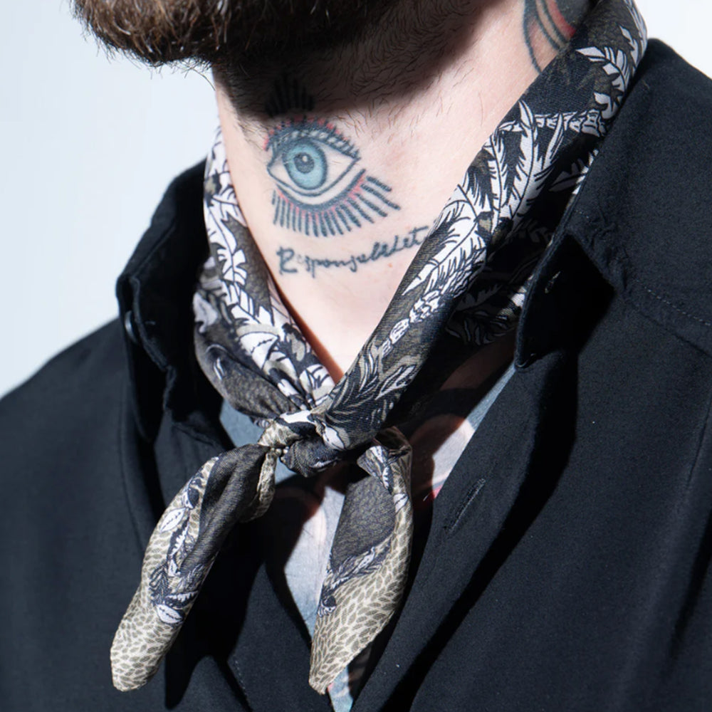 Nahaufnahme eines Mannes mit Tattoos am Hals, der das Mancino Seiden-Foulard "Tropical Green" lässig geknotet über einem schwarzen Hemd trägt.