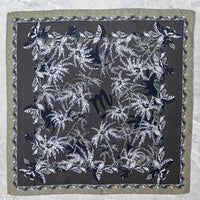 Flach ausgebreitetes Mancino Seiden-Foulard aus 100% Seide. Das Design zeigt ein schwarzes 'm'-Logo im Zentrum, umgeben von detaillierten schwarz-weißen Palmenblättern auf khakigrünem Grund mit gemusterter Bordüre.