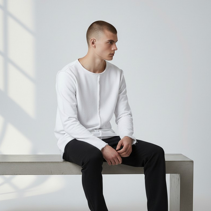 Männliches Model sitzt in dem weißen Langarmshirt Alejandro. Lässiger Look mit sichtbaren, nach außen gekehrten Nähten und offenem Saum.