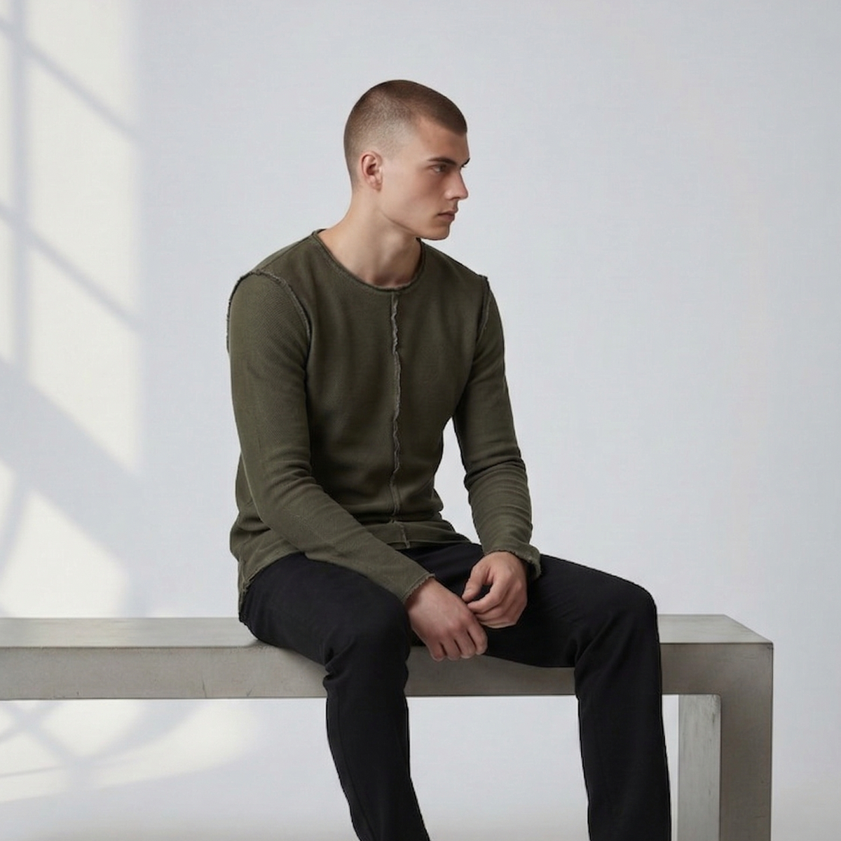 Männliches Model sitzt in dem grünen Langarmshirt Alejandro auf einer Betonbank. Lässiger Look mit sichtbaren, nach außen gekehrten Nähten und offenem Saum.