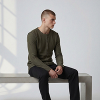 Männliches Model sitzt in dem grünen Langarmshirt Alejandro auf einer Betonbank. Lässiger Look mit sichtbaren, nach außen gekehrten Nähten und offenem Saum.