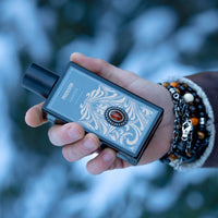 Nahaufnahme einer Hand, die den Flakon des Mancino Amber Eau de Parfum vor einem winterlichen, blau-weiß verschwommenen Hintergrund hält. Das Handgelenk ist mit einer markanten Kombination aus Mancino-Armbändern geschmückt, darunter Tigerauge-Perlen, Silberketten und dunkle Natursteine. Das Detail zeigt das goldene Ornament-Etikett mit dem zentralen, bernsteinfarbenen Schmuckstein auf dem schwarzen Glasflakon.