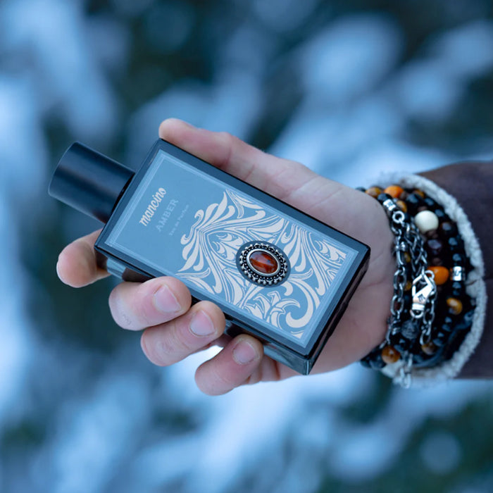 Nahaufnahme einer Hand, die den Flakon des Mancino Amber Eau de Parfum vor einem winterlichen, blau-weiß verschwommenen Hintergrund hält. Das Handgelenk ist mit einer markanten Kombination aus Mancino-Armbändern geschmückt, darunter Tigerauge-Perlen, Silberketten und dunkle Natursteine. Das Detail zeigt das goldene Ornament-Etikett mit dem zentralen, bernsteinfarbenen Schmuckstein auf dem schwarzen Glasflakon.