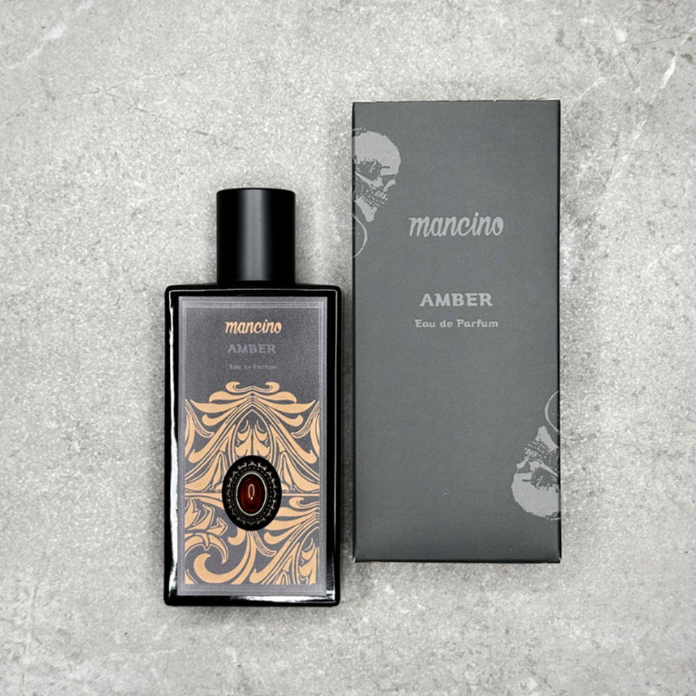 Produktfotografie aus der Vogelperspektive: Der Mancino Amber Eau de Parfum Flakon liegt neben seiner mattschwarzen Umverpackung auf einer grau-strukturierten Steinfläche. Das Etikett besticht durch goldene Verzierungen und einen tiefroten Amber-Schmuckstein. Die schwarze Box zeigt den silbernen „AMBER“-Schriftzug und das charakteristische Totenkopf-Design der Kollektion.