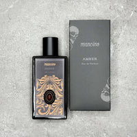 Produktfotografie aus der Vogelperspektive: Der Mancino Amber Eau de Parfum Flakon liegt neben seiner mattschwarzen Umverpackung auf einer grau-strukturierten Steinfläche. Das Etikett besticht durch goldene Verzierungen und einen tiefroten Amber-Schmuckstein. Die schwarze Box zeigt den silbernen „AMBER“-Schriftzug und das charakteristische Totenkopf-Design der Kollektion.
