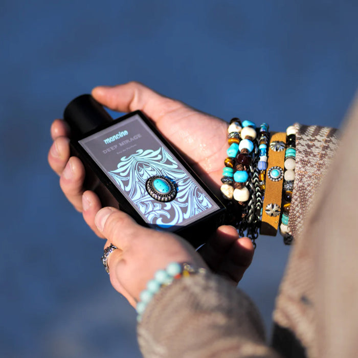 Nahaufnahme von zwei Händen, die den Flakon des Mancino Deep Mirage Eau de Parfum über einem glitzernden, türkisfarbenen Pool halten. Die Handgelenke sind mit einer Vielzahl von Mancino-Accessoires geschmückt, darunter Perlenarmbänder mit Türkis-Steinen, Gliederketten und Lederbänder. Das Detail zeigt das aufwendige Ornament-Etikett mit dem passenden türkisfarbenen Schmuckstein auf dem dunklen Glasflakon.