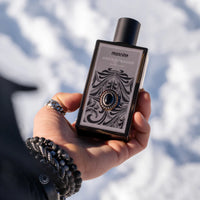 Nahaufnahme einer Hand, die das Mancino Onyx Tobacco Eau de Parfum vor einem unscharfen, sonnigen Schneehintergrund hält. Die Hand trägt mehrere Mancino-Schmuckstücke, darunter ein Gliederring und zwei Obsidian-Perlenarmbänder. Die Detailansicht zeigt das aufwendige, ornamentale Etikett des dunklen Glasflakons mit dem zentralen Onyx-Stein-Detail.