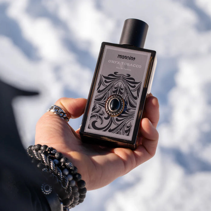 Nahaufnahme einer Hand, die das Mancino Onyx Tobacco Eau de Parfum vor einem unscharfen, sonnigen Schneehintergrund hält. Die Hand trägt mehrere Mancino-Schmuckstücke, darunter ein Gliederring und zwei Obsidian-Perlenarmbänder. Die Detailansicht zeigt das aufwendige, ornamentale Etikett des dunklen Glasflakons mit dem zentralen Onyx-Stein-Detail.
