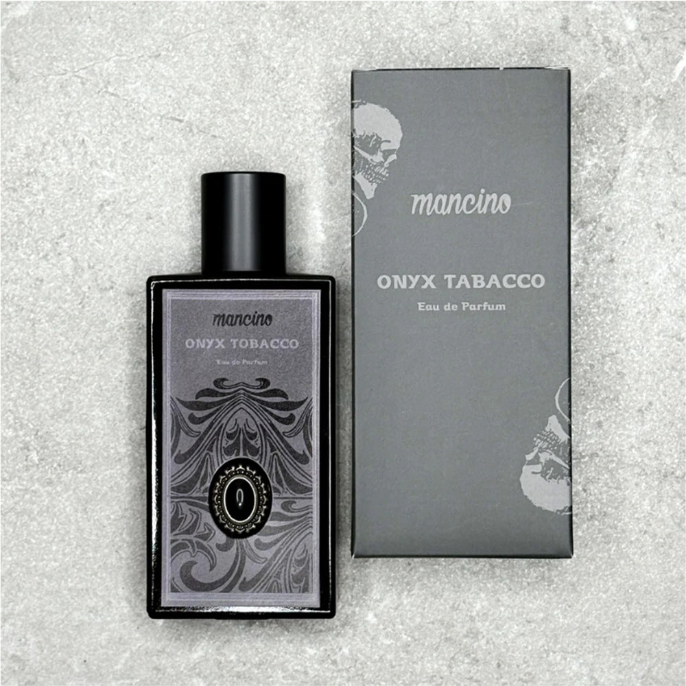 Flatlay-Produktfotografie des Mancino Onyx Tobacco Eau de Parfum neben seiner mattschwarzen Umverpackung auf einer grauen strukturierten Steinoberfläche. Der dunkle Glasflakon ist mit dem detaillierten silbernen Etikett und dem Onyx-Zierstein versehen. Die schwarze Box zeigt das Mancino-Logo und den Duftnamen in Silber sowie ein dezentes Totenkopf-Motiv im Hintergrund.