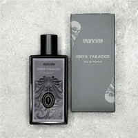 Flatlay-Produktfotografie des Mancino Onyx Tobacco Eau de Parfum neben seiner mattschwarzen Umverpackung auf einer grauen strukturierten Steinoberfläche. Der dunkle Glasflakon ist mit dem detaillierten silbernen Etikett und dem Onyx-Zierstein versehen. Die schwarze Box zeigt das Mancino-Logo und den Duftnamen in Silber sowie ein dezentes Totenkopf-Motiv im Hintergrund.