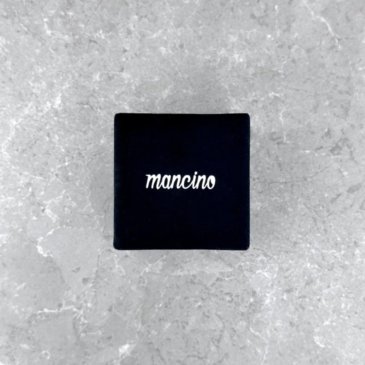 Geschlossene, mattschwarze Mancino Ringbox mit weißem Marken-Schriftzug, edel platziert auf einem grauen Steinuntergrund.