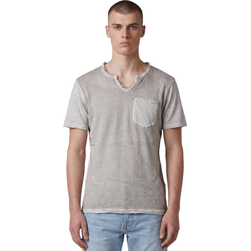 Frontansicht eines männlichen Models, das das beigefarbene T-Shirt mit V-Ausschnitt trägt. Deutlich erkennbar sind die strukturierte Slub-Baumwolle, die offenkantigen Raw-Edge-Details an Kragen, Ärmeln und Bund sowie die aufgesetzte Brusttasche. Das Shirt wird lässig zu einer hellblauen Jeans präsentiert.