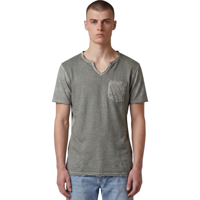 Frontansicht eines männlichen Models, das das salbeigrüne T-Shirt mit V-Ausschnitt trägt. Deutlich erkennbar sind die strukturierte Slub-Baumwolle, die offenkantigen Raw-Edge-Details an Kragen, Ärmeln und Bund sowie die aufgesetzte Brusttasche. Das Shirt wird lässig zu einer hellblauen Jeans.